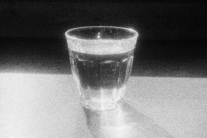 A glass of water on a table, its bright rim spilling light into the dark behind it